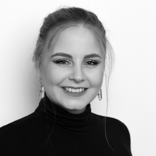 Porträt von Judith Sandmann. Sie trägt ein schwarzes Oberteil mit Rollkragen und auffällige Ohrringe. Ihre blonden Haare sind hochgesteckt, einzelne Strähnen umrahmen das Gesicht. Sie lächelt freundlich in die Kamera, vor einem hellen Hintergrund.