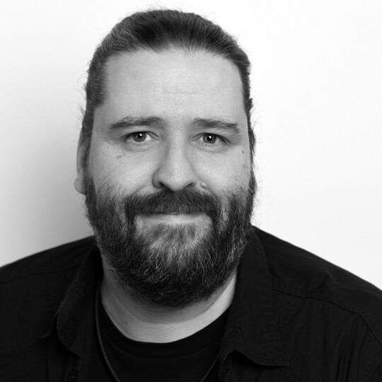 Porträt von Mario Schnabl. Er trägt ein schwarzes Hemd und hat einen Vollbart sowie lange Haare, die nach hinten gebunden sind. Er blickt freundlich in die Kamera, vor einem hellen Hintergrund.