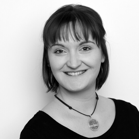 Porträt von Mirijam Fein-Scheunert. Sie trägt ein dunkles Oberteil und eine Halskette mit rundem Anhänger. Sie hat rötlich-braunes, schulterlanges Haar mit Pony und lächelt in die Kamera. Der Hintergrund ist hell.