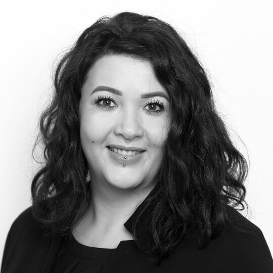 Porträt von Angelique Lobjinski. Sie hat schulterlanges, dunkel gelocktes Haar und trägt ein schwarzes Oberteil. Sie lächelt freundlich in die Kamera. Sichtbar sind ein Nasen- und ein Lippenpiercing. Der Hintergrund ist hell.