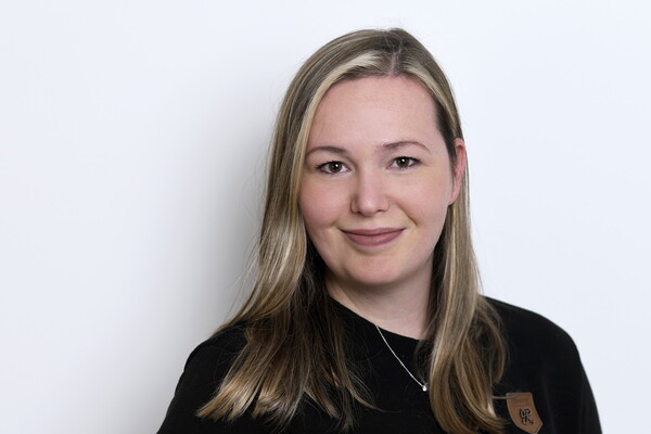 Porträt von Lisa Sittig. Sie trägt ein dunkles Oberteil mit einem kleinen Leder-Emblem und eine schlichte Halskette. Ihr blondes, glattes Haar ist offen getragen. Sie lächelt leicht in die Kamera, vor einem hellen Hintergrund.