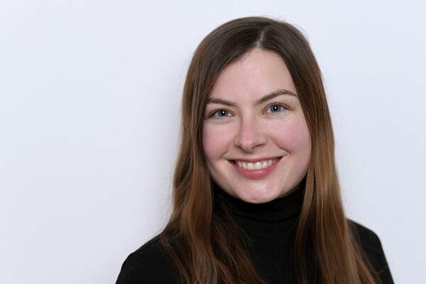 Porträt von Romy Kleinert. Sie hat langes, glattes, braunes Haar und trägt ein schwarzes Oberteil mit Rollkragen. Sie lächelt freundlich in die Kamera, vor einem hellen Hintergrund.
