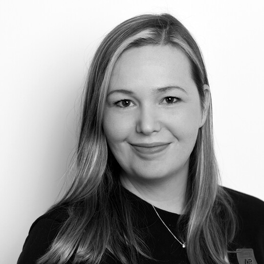 Porträt von Lisa Sittig. Sie trägt ein dunkles Oberteil mit einem kleinen Leder-Emblem und eine schlichte Halskette. Ihr blondes, glattes Haar ist offen getragen. Sie lächelt leicht in die Kamera, vor einem hellen Hintergrund.