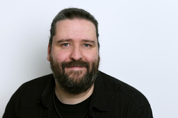 Porträt von Mario Schnabl. Er trägt ein schwarzes Hemd und hat einen Vollbart sowie lange Haare, die nach hinten gebunden sind. Er blickt freundlich in die Kamera, vor einem hellen Hintergrund.