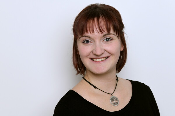 Porträt von Mirijam Fein-Scheunert. Sie trägt ein dunkles Oberteil und eine Halskette mit rundem Anhänger. Sie hat rötlich-braunes, schulterlanges Haar mit Pony und lächelt in die Kamera. Der Hintergrund ist hell.