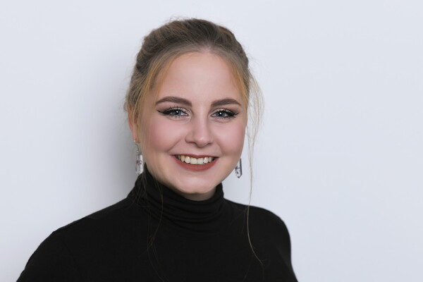 Porträt von Judith Sandmann. Sie trägt ein schwarzes Oberteil mit Rollkragen und auffällige Ohrringe. Ihre blonden Haare sind hochgesteckt, einzelne Strähnen umrahmen das Gesicht. Sie lächelt freundlich in die Kamera, vor einem hellen Hintergrund.